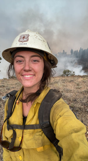 Sahale DelCampo in firefighting gear with a helmet and yellow jacket, standing in front of smoke and flames.