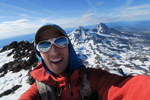 Kyle Bingham taking a selfie on a snowy mountain peak with a clear blue sky.